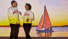 Two alums smiling with sail boat on Lake Mendota in background.