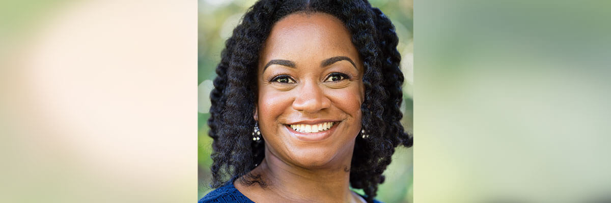 A photograph of professor Raquel Kennon set against a blurred green background. She is smiling, her hair is down, and she is wearing a royal blue top and dangling earrings with white stones.