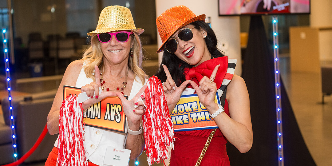 Two alumnae pose for a silly photo with spirited props and sequined hats.